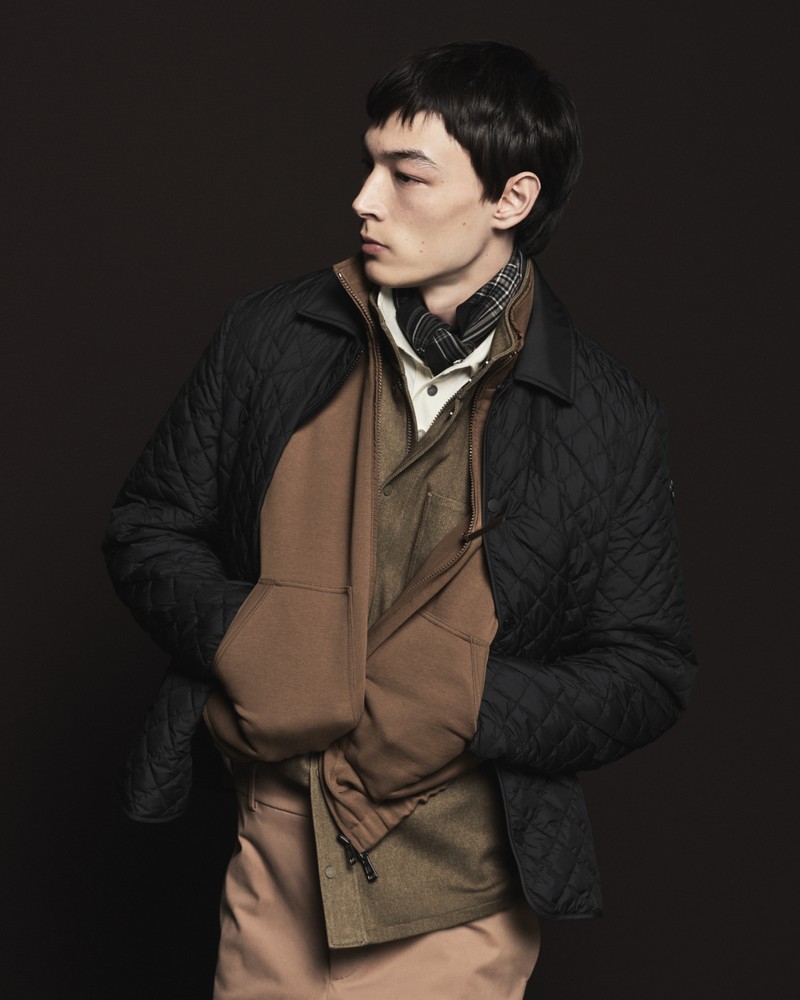 Piumino trapuntato scuro Moncler Pre-Fall 2026 su sciarpa, felpa, camicia bianca e pantaloni cammello.