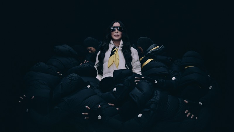 Moncler Re/Icons 2026，身穿白色羽绒服、系黄色丝巾的Cher，被身穿黑色羽绒服的人物环绕。