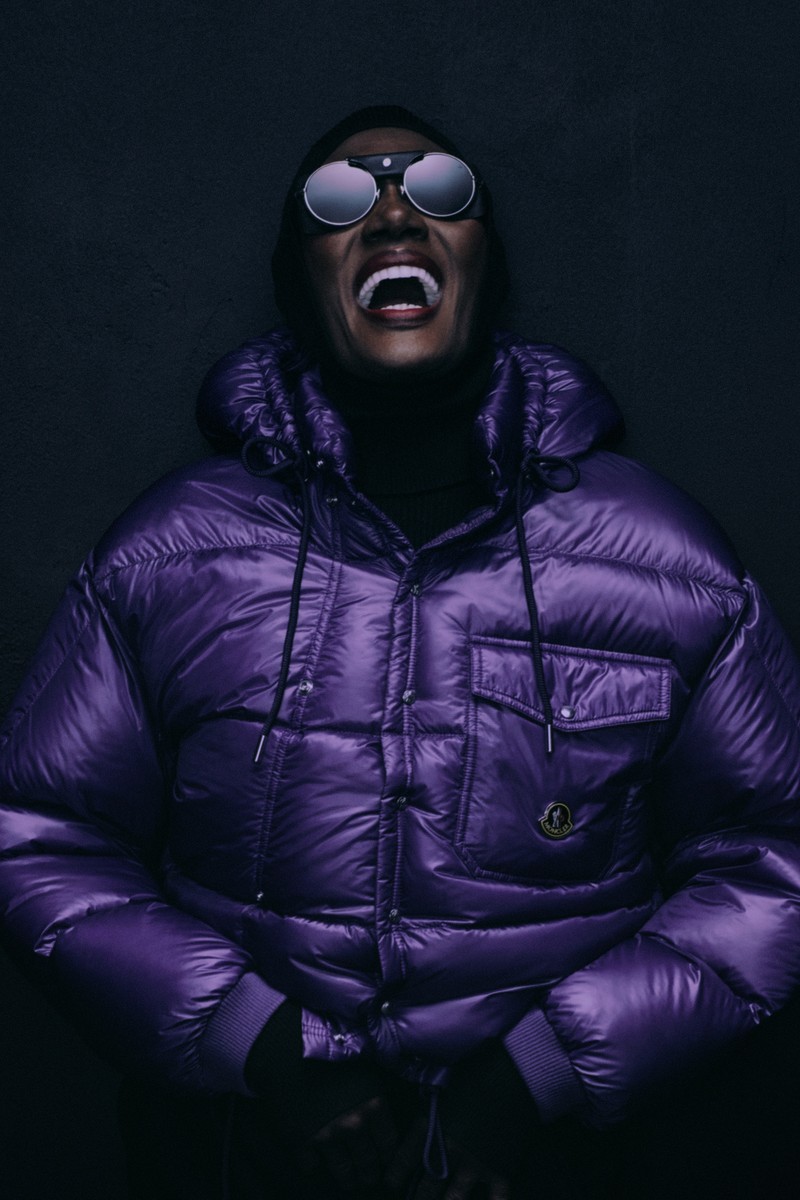 Moncler Re/Icons 2026, Pioneers in Moncler: Grace Jones in violettem Daunenmantel inmitten von Figuren in dunklen Anzügen.