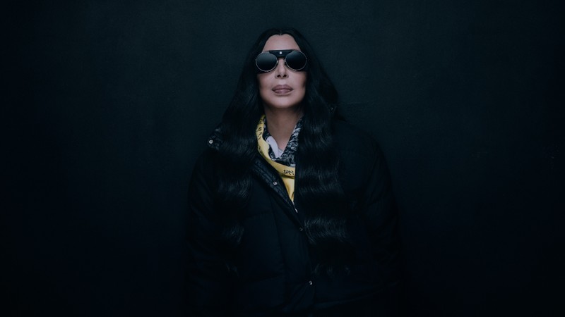 Moncler Re/Icons 2026, portrait of Cher in round sunglasses, a black down jacket and a yellow scarf. 