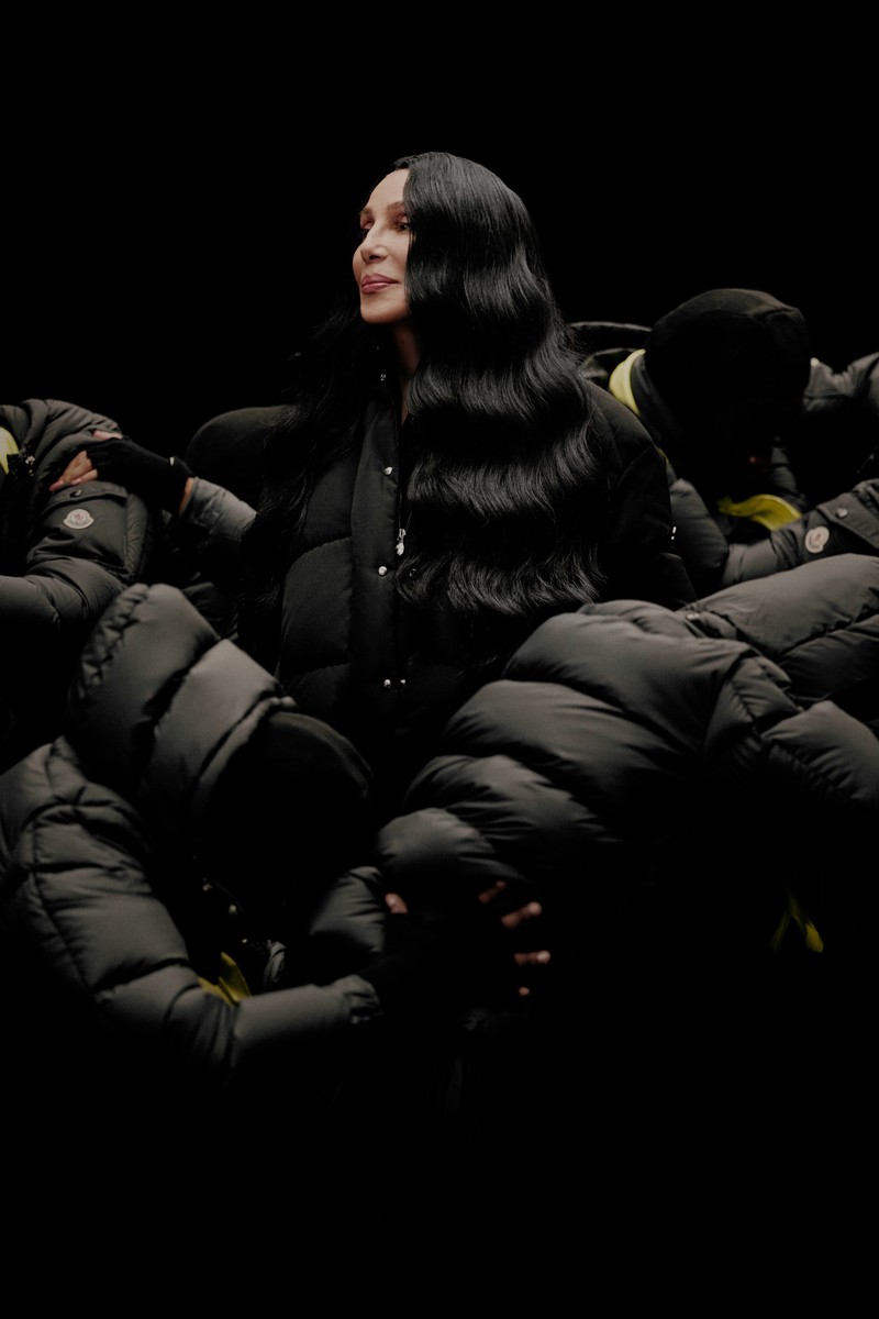 Moncler Re/Icons 2026, behind the scenes in black and white: Cher on set with a cast in black down jackets. 