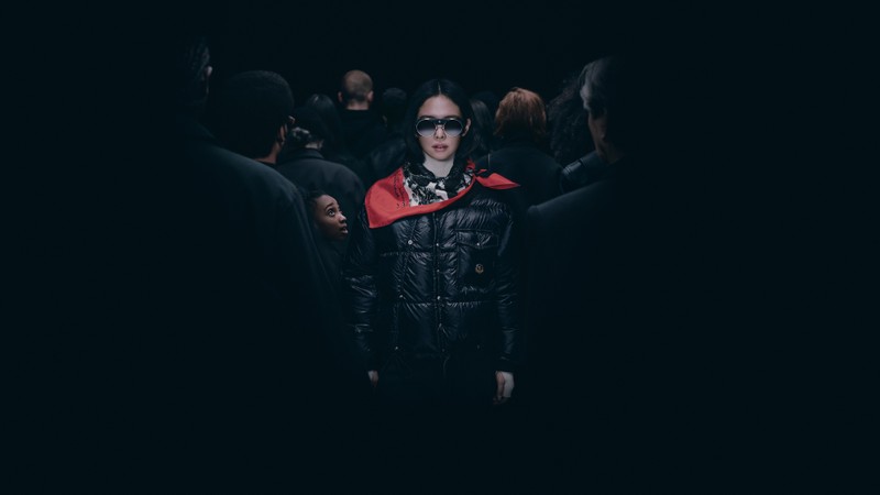 Moncler Re/Icons 2026, Jennie in schwarzer Daunenjacke und rotem Tuch, inmitten einer schwarz gekleideten Menge.