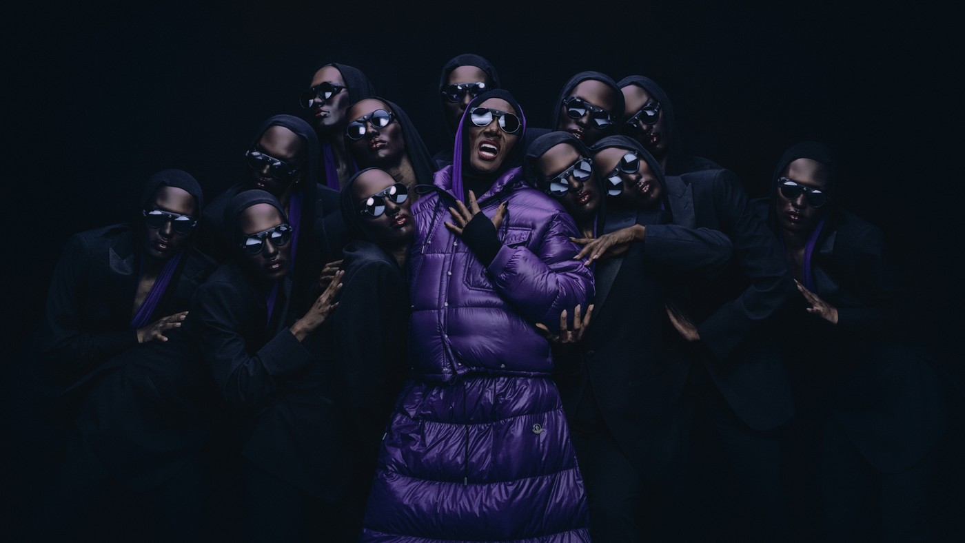 Grace Jones in a purple Moncler down coat surrounded by similarly styled figures in dark suits and sunglasses