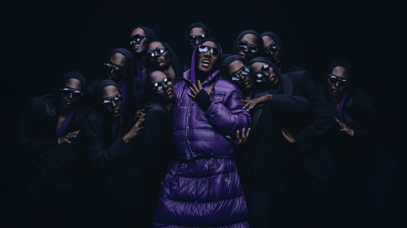 Moncler Re/Icons 2026、サングラスとダークスーツの人々に囲まれた、パープルのロングダウンコート姿のGrace Jones。