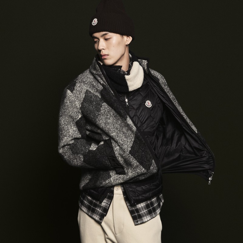 Moncler メンズ。ダークブラウンのニット帽、グレーのパッチワークカーディガン、ブラックのキルティングダウンジャケット、チェック柄シャツとベージュのパンツ。 