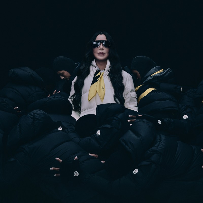 Moncler Re/Icons 2026, Pioneers in Moncler: Cher con plumífero blanco y pañuelo amarillo entre figuras con plumíferos negros.