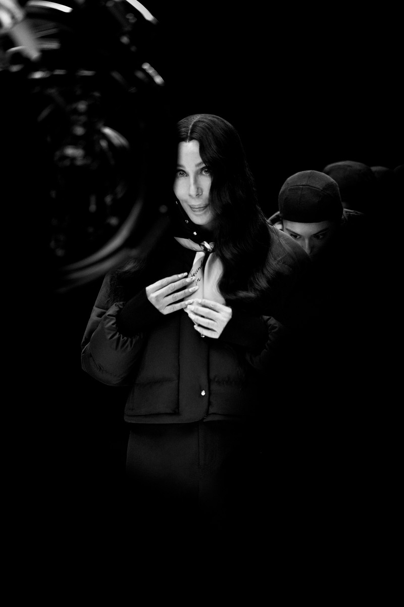 Moncler Re/Icons 2026, behind the scenes in black and white: Cher on set with a cast in black down jackets. 