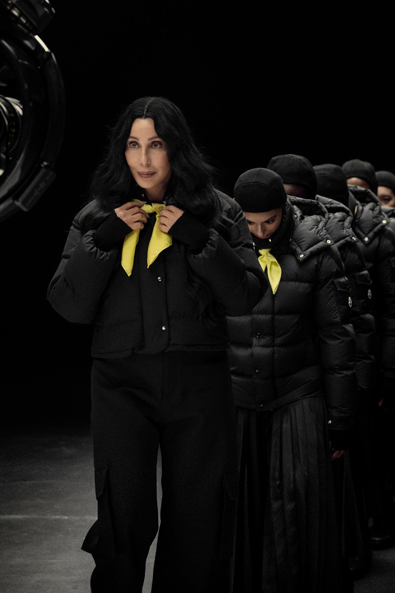 Moncler Re/Icons 2026, behind the scenes in black and white: Cher on set with a cast in black down jackets. 