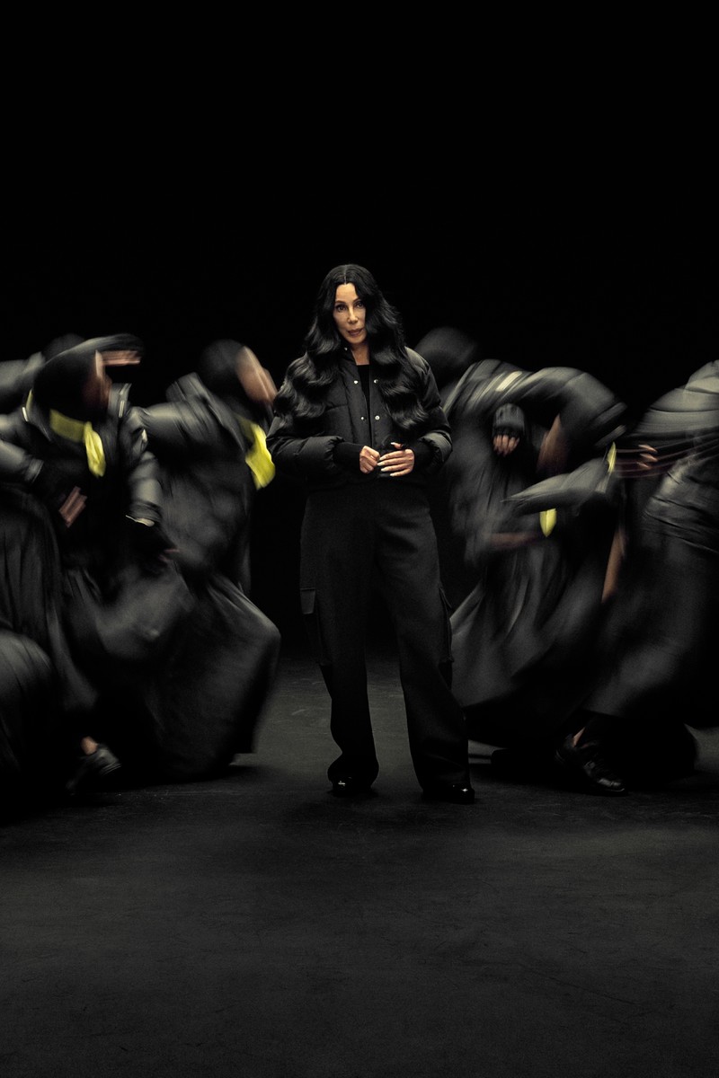 Moncler Re/Icons 2026, behind the scenes in black and white: Cher on set with a cast in black down jackets. 