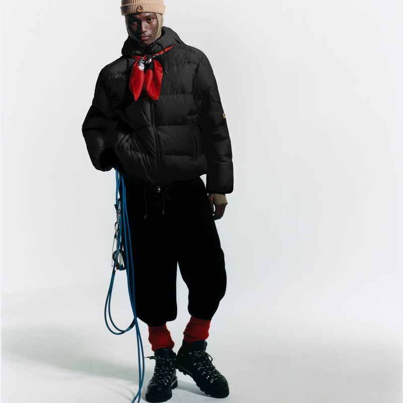 Moncler Re-Icons、Himalaya。メンズの黒のダウンジャケット、赤いスカーフとベージュの帽子、ハイキングブーツと青いロープ。